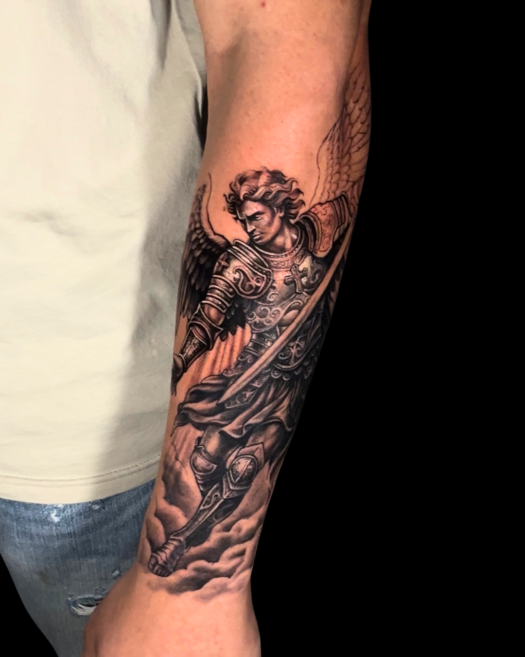 Tatuagem religiosa realista no antebraço com arcanjo, asas, armadura e espada em preto e cinza.