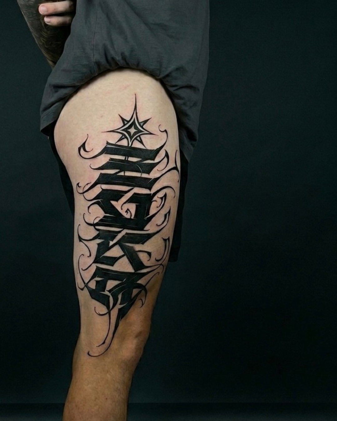 Tatuagem personalizada de lettering na coxa com letras pretas e detalhes ornamentais.