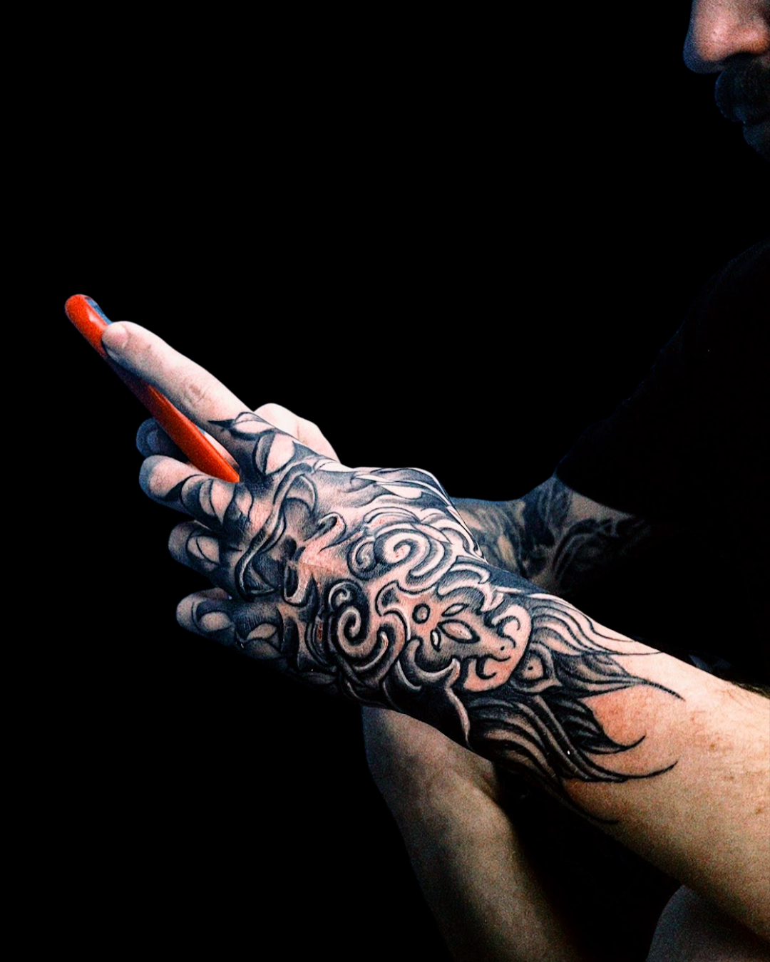 Tatuagem na mão (hand tattoo) em estilo oriental de um Foo Dog preto e cinza. A arte oriental cobre as costas da mão e os dedos com linhas fluidas, espirais e sombreamento, criando um design decorativo e único.