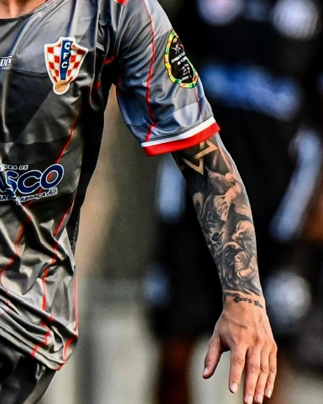 Tatuagem de braço fechado (full sleeve) de um jogador de futebol, no estilo realismo preto e cinza. A arte inclui figuras de anjos, rostos, pombas e uma escrita no pulso, compondo um projeto de tattoo religiosa e geométrica.