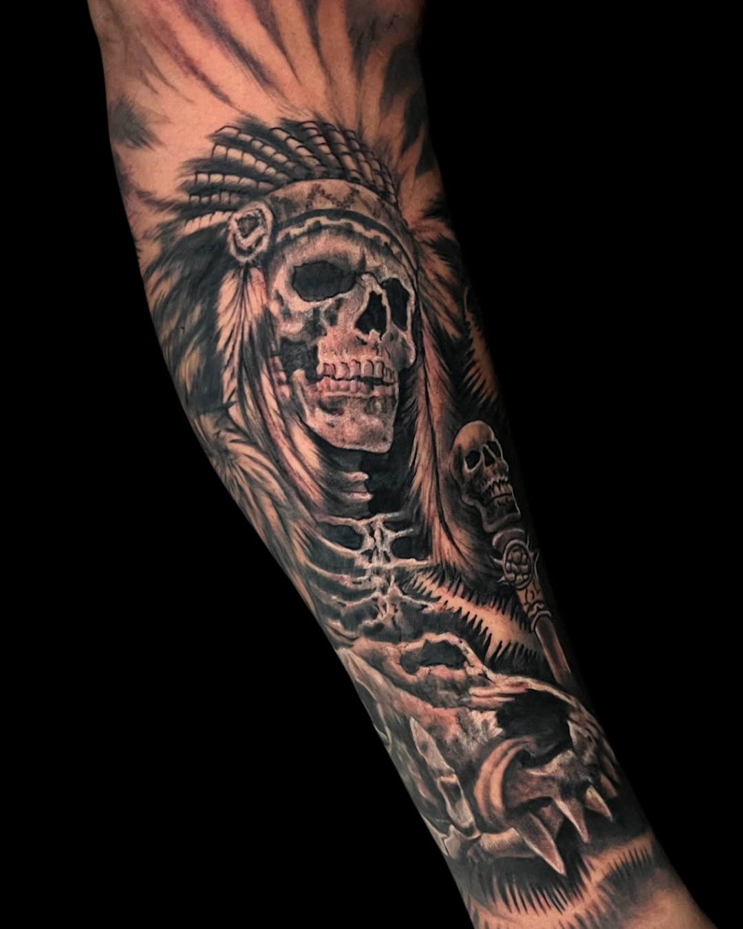 Tatuagem de antebraço em realismo preto e cinza, mostrando uma caveira com um cocar indígena americano detalhado. A composição inclui um esqueleto e outras caveiras, criando uma tattoo com tema de guerreiro e mortalidade.