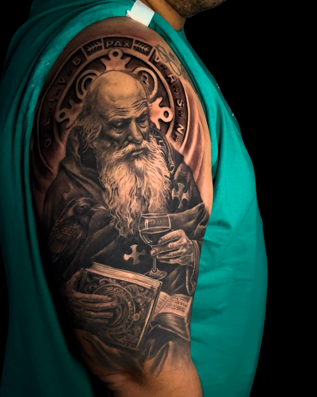 Tatuagem realista religiosa no braço em preto e cinza com santo, livro e cálice.